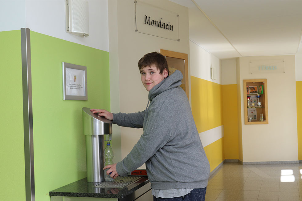 Ein junger Patient steht an einem Wasserspender und hält eine leere Trinkflasche darunter. Ein junger Patient steht an einem Wasserspender und hält eine leere Trinkflasche darunter.