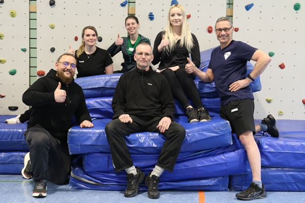 Drei Frauen und drei Männer vor einer Sportmatte Drei Frauen und drei Männer vor einer Sportmatte