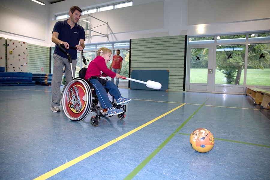Ein Junge im Rollstuhl spielt mit Unterstützung eines Therapeuten in der Sporthalle. Ein Junge im Rollstuhl spielt mit Unterstützung eines Therapeuten in der Sporthalle.