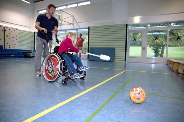 Ein Junge im Rollstuhl spielt mit Unterstützung eines Therapeuten in der Sporthalle.